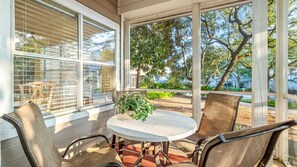 Une terrasse couverte avec une table ronde et deux chaises, de grandes fenêtres et une vue sur les arbres et une maison.