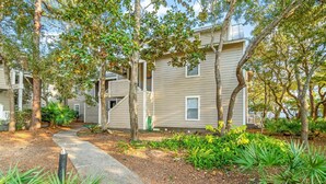 A residential building with multiple floors, a covered outdoor area, and a pathway surrounded by trees and plants. - Hidden Dunes Beach Cottage 178 - Charming Beach Cottage! (Miramar Beach)