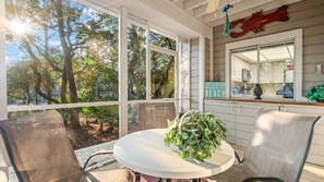 Un espace extérieur aménagé, abrité, avec une table et des chaises, une plante décorative, et une vue sur des arbres et un panneau indiquant la direction de la plage.