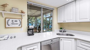 A kitchen with white cabinets, a Bosch dishwasher, a coffee maker, and a window with a view of trees and a building. - Hidden Dunes Beach Cottage 178 - Charming Beach Cottage! (Miramar Beach)
