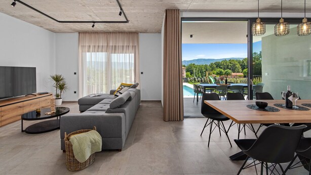 Un salon moderne avec un canapé, un coin repas comprenant une table et des chaises, et une grande fenêtre donnant sur une piscine et des montagnes.