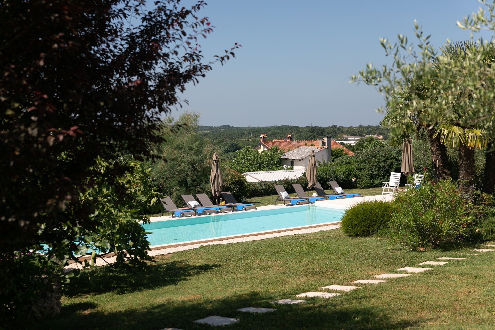 A pool area with lounge chairs and umbrellas, surrounded by greenery and buildings in the background.