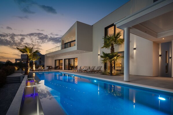 Un espace moderne aménagé au bord de la piscine, comprenant une piscine, des chaises longues et un bâtiment à deux étages doté de grandes fenêtres.