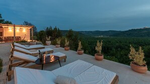 Um terraço com cadeiras de descanso, uma mesa com garrafa e copos, e plantas em vasos.