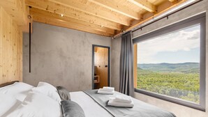 Quarto moderno com uma cama grande, roupa de cama cinzenta e vista para uma paisagem arborizada através de uma janela ampla.