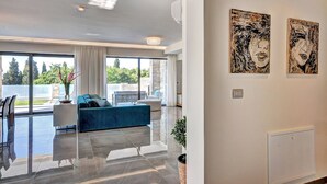 Un salon moderne avec un canapé bleu, un coin repas comprenant une table et des chaises, de grandes fenêtres donnant sur un paysage verdoyant, et deux tableaux abstraits accrochés au mur.