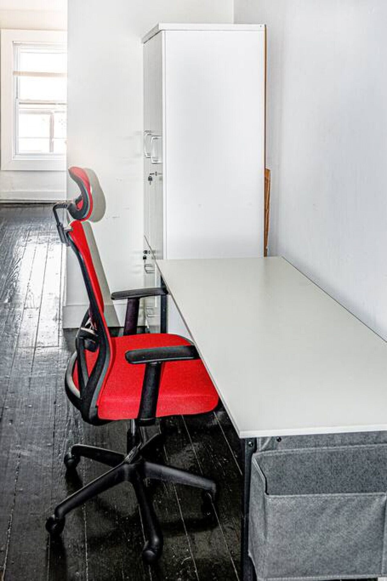 A small office space with a white desk, a red office chair, and a grey storage unit.