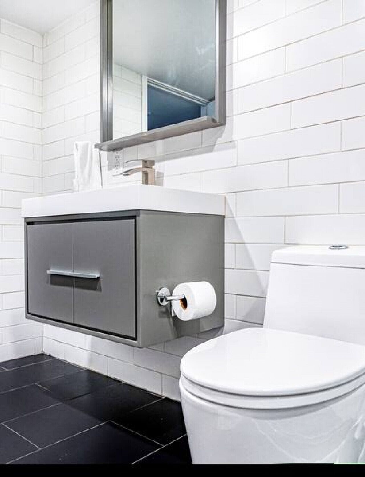 A modern bathroom with a toilet, a grey cabinet with a toilet paper holder, and a mirror above the sink.