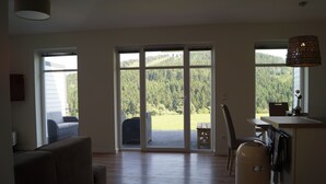 Een ruime woonkamer met een grote schuifpui naar een terras, een bank, een prullenbak en een hanglamp.