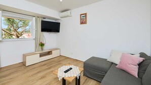 Een moderne woonkamer met een grijze bank, een klein houten tafeltje met een afstandsbediening, een aan de muur bevestigde televisie en een raam met uitzicht op bomen en gebouwen.