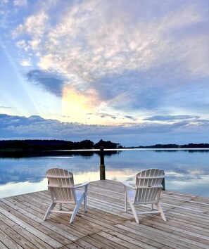 En træterrasse med to hvide Adirondack-stole, der vender ud mod en rolig sø, med en solnedgang i baggrunden.