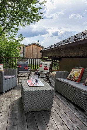 Una terraza al aire libre con muebles de mimbre, una mesa con vasos y una botella, y un edificio de madera al fondo.