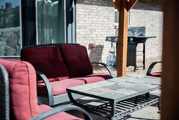 A patio area with a red cushioned sofa, a metal table, and a grill in the background. - Craftsman Charm Comfort 3BDR (El Paso)