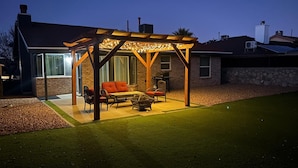 Un espacio al aire libre techado con mesa y sillas, una chimenea y una pérgola con luces colgantes.