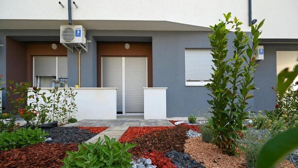 Un bâtiment moderne à la façade grise, doté de deux portes fermées et d’un petit jardin agrémenté de diverses plantes et d’un chemin de gravier.