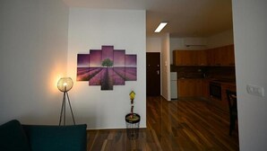 Un salon moderne avec un canapé d’angle, une lampe sur pied et un tableau accroché au mur. On aperçoit une petite table d’appoint sur laquelle est posé un vase. Le salon donne sur une cuisine avec des meubles en bois et un réfrigérateur.