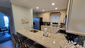 A kitchen with a granite countertop, stainless steel sink, and a built-in oven. There are bar stools and a hanging light fixture. - RENOVATED LUXURY BEACH VILLA,Ocean view,3 bdrms each with smart tv and king bed (Hilton Head Island)