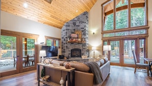 A cozy living room with a stone fireplace, wooden ceiling, and large windows offering a view of the outdoors.