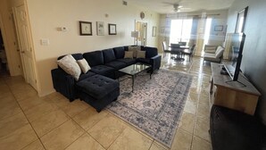 A spacious living room with a sectional sofa, a coffee table, and a television. There is a dining area with a table and chairs, and a ceiling fan.