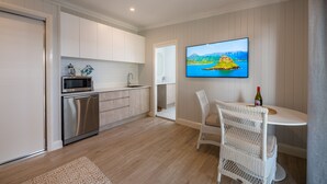 A modern kitchen with white cabinets, a stainless steel dishwasher, and a microwave. There is a dining area with a white wicker chair and a table, and a flat-screen TV mounted on the wall displaying a scenic landscape. - Oasis on the Esplanade (Manly)