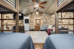 A rustic room with a stone wall, a ceiling fan, a wooden door, and a bed with a patterned bedspread. - Cypress Star on the Medina | 4BR with Pool & River (Bandera)