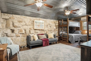 A rustic interior with a stone wall, a grey sofa with cushions, a wooden table, and a ceiling fan.