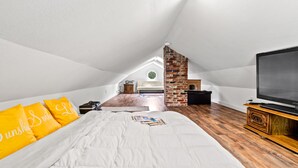 Ein Schlafzimmer mit erhöhter Decke, einem Bett mit weißer Bettwäsche und gelben Kissen, einem Kamin aus Ziegelsteinen, einem Fernseher auf einem Holzboden und einem Holzboden.