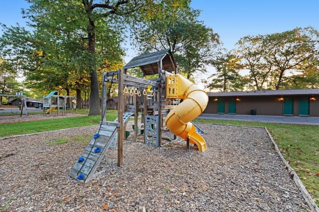 A playground with a slide and climbing structures, surrounded by trees and a building in the background.