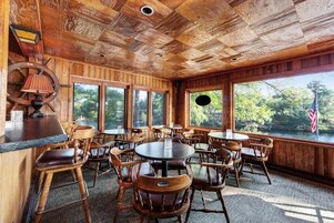 A rustic wooden interior with round tables and chairs, large windows offering views of the outdoors, and a bar area with a lamp and American flag.