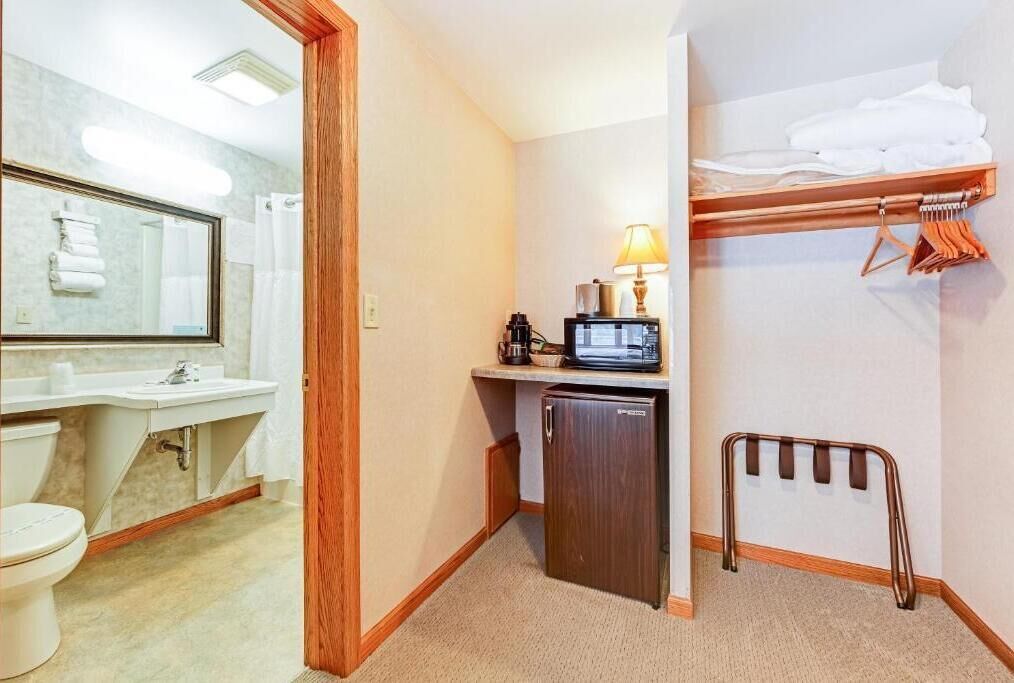 A compact hotel bathroom with a sink, toilet, mirror, and a small refrigerator.