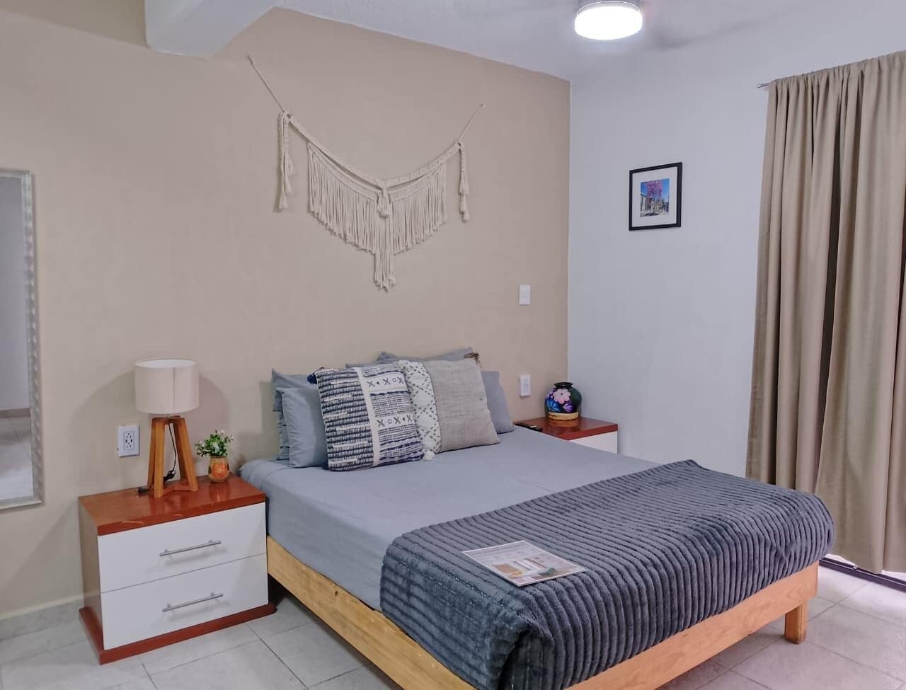 A bedroom with a bed, bedside tables, a wall hanging, a picture, and a window with curtains.