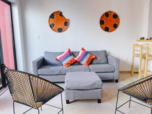 Una sala de estar con un sofá gris, dos almohadas coloridas, un reposapiés gris y dos sillas de mimbre negras. Hay dos adornos en la pared que recuerdan a mariquitas.