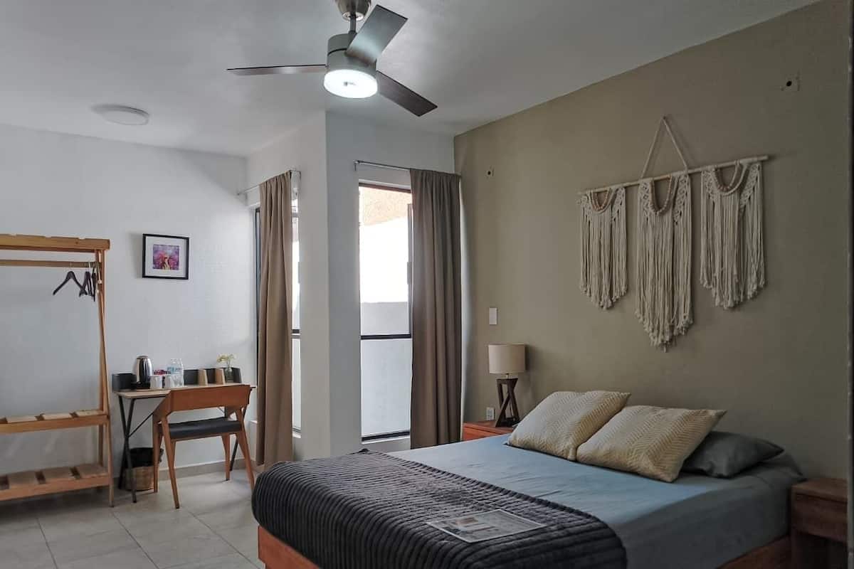 A bedroom with a bed, a desk, a chair, a ceiling fan, and a macrame wall hanging.