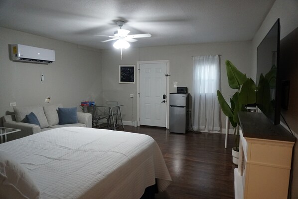 A hotel room with a bed, a desk, a chair, a refrigerator, a ceiling fan, and a large window with curtains.