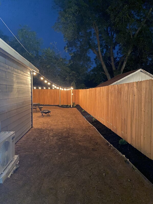 A backyard with a wooden fence, a patio area with a table and chairs, and string lights.