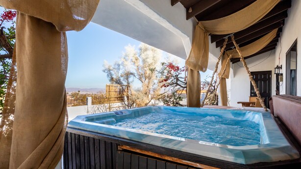 Un bain à remous doté d’une terrasse en bois, de rideaux beiges et offrant une vue sur les arbres et un paysage lointain.