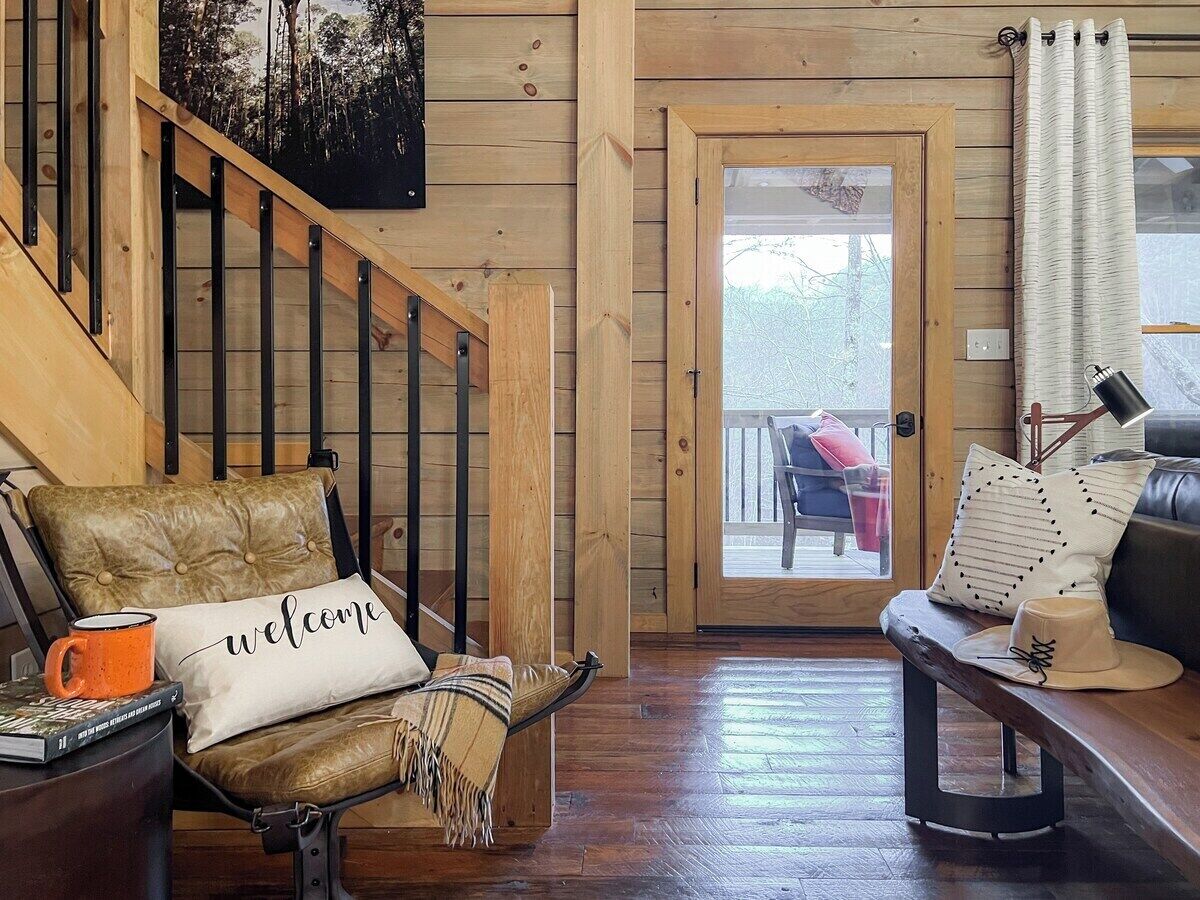 Interior de madera con una escalera, un banco acolchado con un cojín que dice 'welcome', una mesa de centro y vistas a una terraza exterior a través de una puerta de cristal.