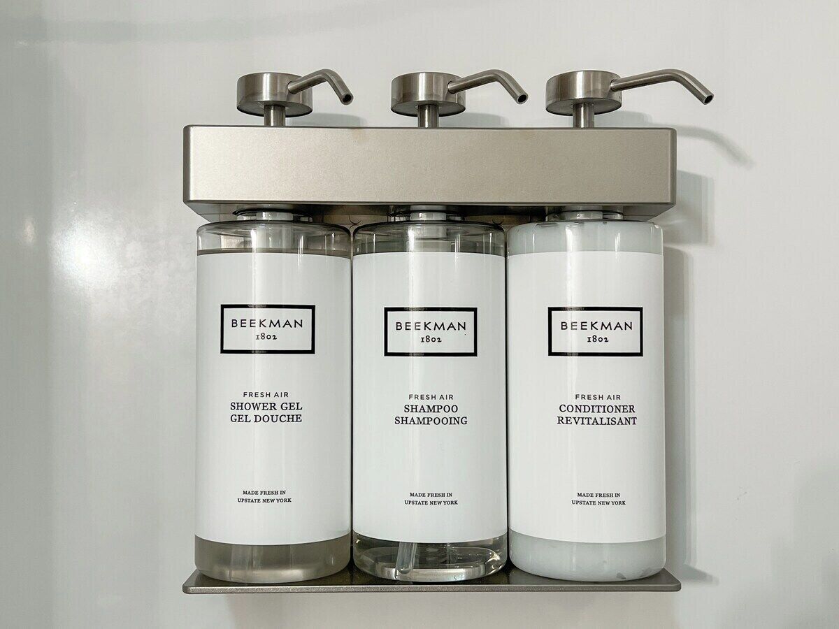 Kolme Beekman-tuotepulloa – suihkugeeli, shampoo ja hoitoaine – on asetettu esille metalliselle hyllylle, jonka yläpuolella on hana.