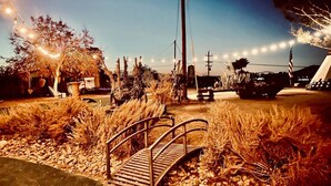 Un entorno rústico al aire libre con un puente de madera, luces colgantes y diversas plantas.