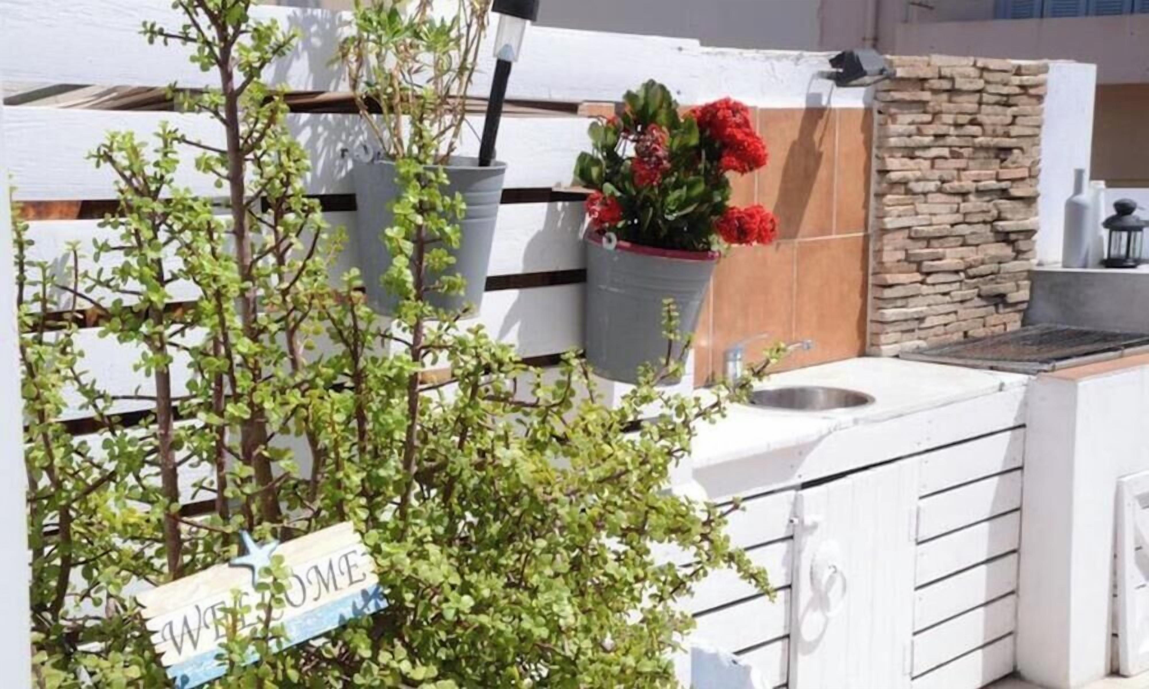 Un espace cuisine extérieur blanc, agrémenté d’une pancarte « BIENVENUE », de plantes en pot et d’un mur en pierre.