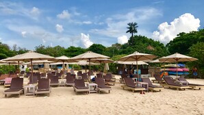 A beach with rows of sun loungers and umbrellas, a palm tree, and a surfboard rack. - "nusa Dua Luxury 2 Bedroom Apartment" (Nusa Dua)