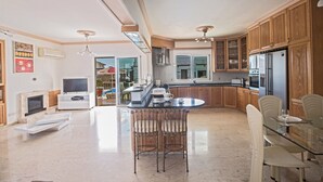 A modern kitchen with wooden cabinets, a central island, and a dining area with a glass table and chairs. - Papas Family Villa With Private Pool Wheelchair Accessible (Paralimni)