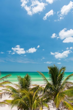 A beach with clear turquoise water, white sand, and palm trees under a blue sky with scattered clouds.