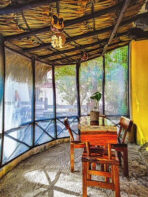 A room with a wooden table, chairs, a window with a view of greenery, and a thatched roof.