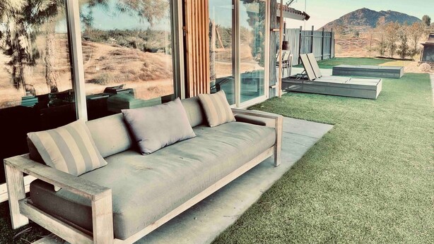 A modern outdoor seating area with a wooden bench and cushions, large windows offering a view of a grassy landscape and mountains. - Love Casa Pool Fire Pit Close To Wineries (Temecula)