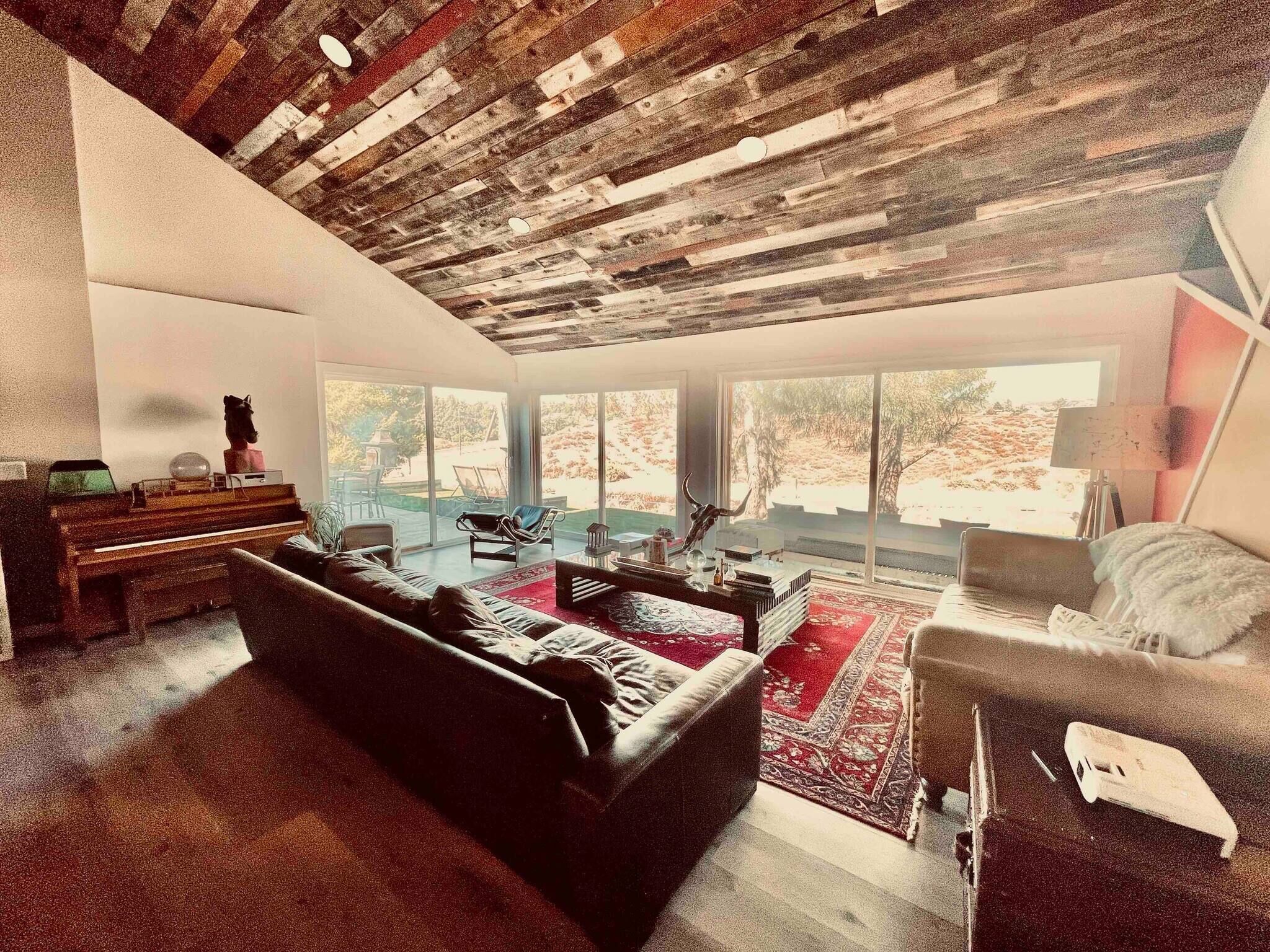Amplia sala de estar con un sofá negro, una mesa de centro y un piano. Grandes ventanales ofrecen vistas al exterior.