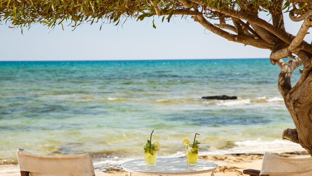 A beach scene with a round table and two chairs under a tree, overlooking the ocean. - Sanders Crystal 2 - Cute 1-bdr Apt With Shared Rooftop (Limassol)