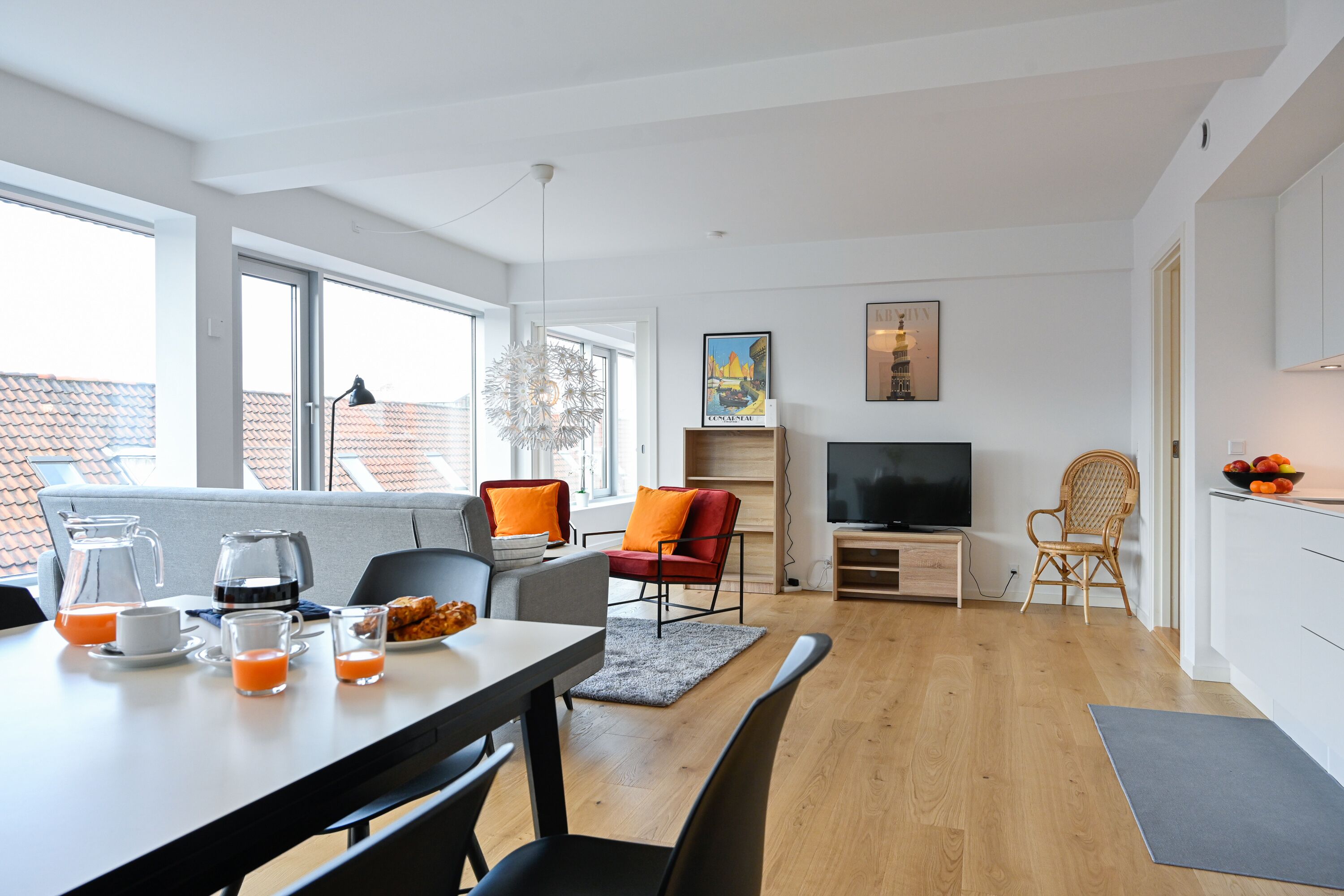 A modern living room with a dining area, large windows, a flat-screen TV, and a kitchen area with fruit on the counter.