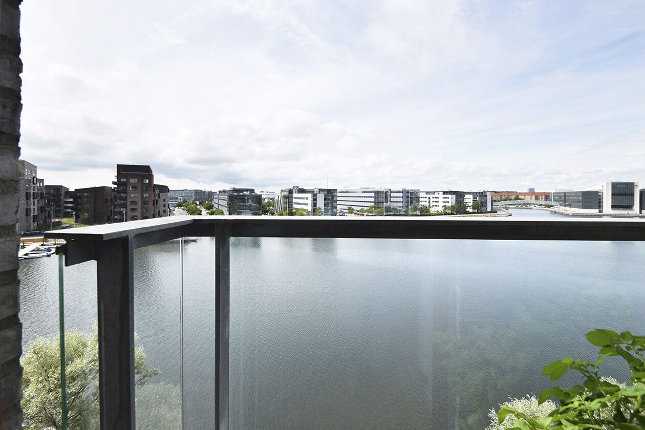 Un edificio moderno con balcón de vidrio que da a una masa de agua y un paisaje urbano.