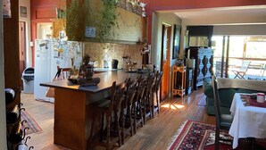 A cozy kitchen with wooden cabinets, a built-in island, and a variety of plants. There is a dining area with a table and chairs, and a view of the outdoors through large windows. - Room-living In Camps Bay Blue (Cape Town)